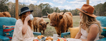 Two women sitting outdoors with Highland cows, under a sign for 'The Cowabunga by Havenfield'.